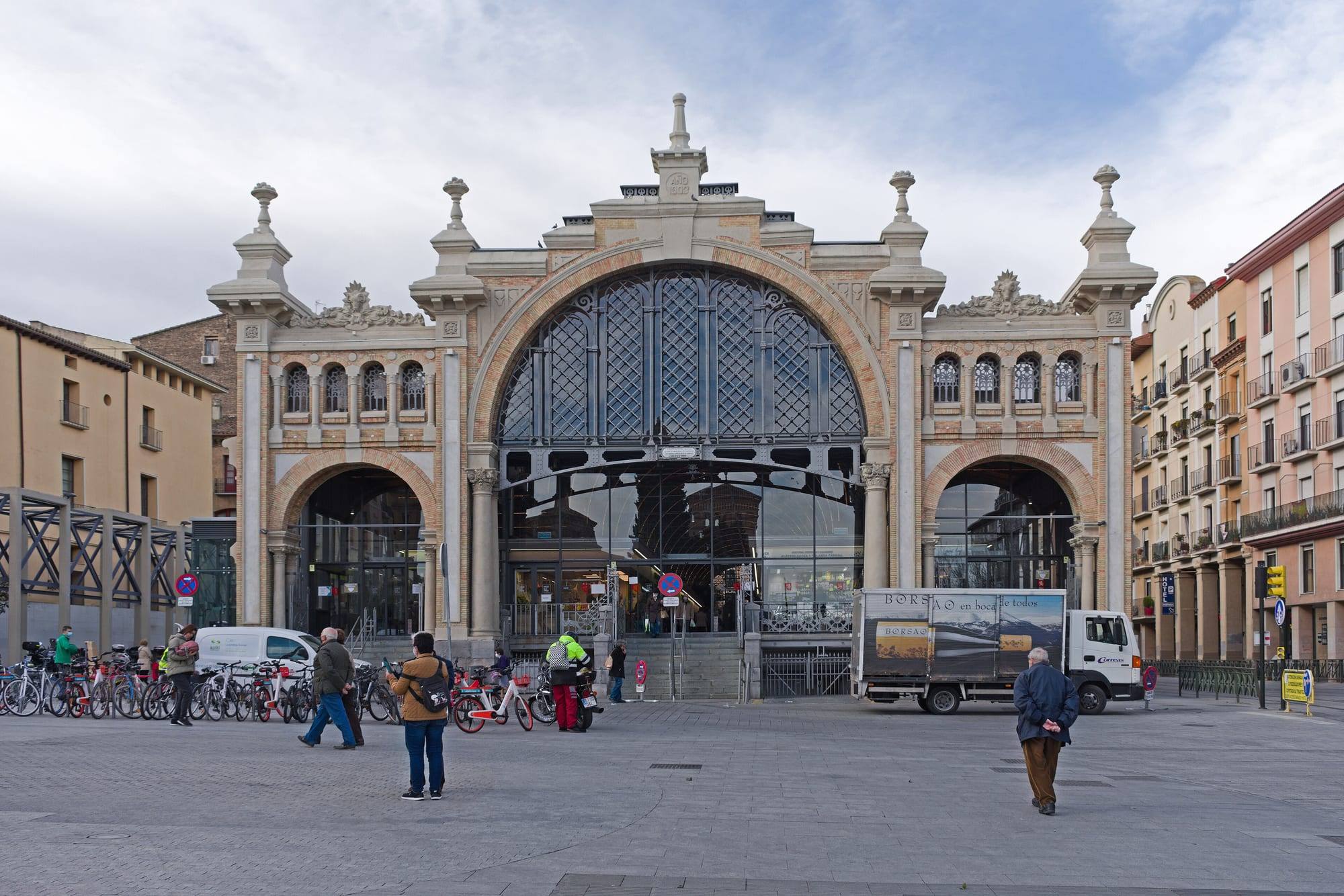 The Mercado Central of Zaragoza, a historic market hall with a large arched iron-and-glass entrance, with shoppers and parked bicycles on the square in front