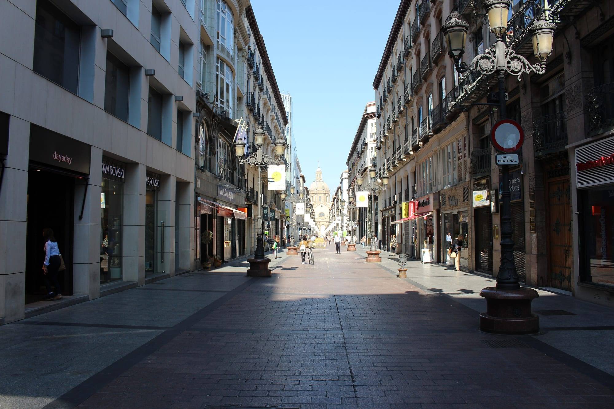 Calle de Alfonso I, a pedestrian street of Zaragoza's old town lined with shops, ornate buildings and cast-iron street lamps, with a dome of the Basílica del Pilar at its end